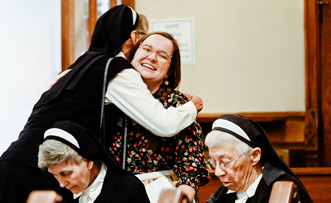 two nuns with heads bowed praying while another nun hugs a woman in a flower print dress