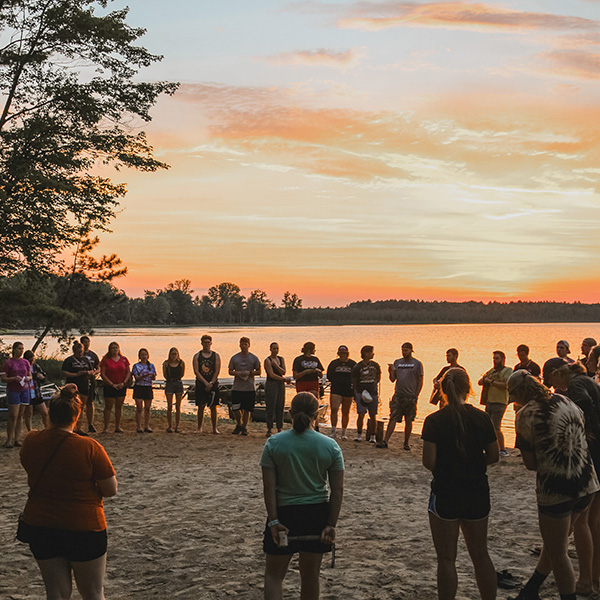 lots of young adults standing in a circle on a beach in front of a beautiful sunset