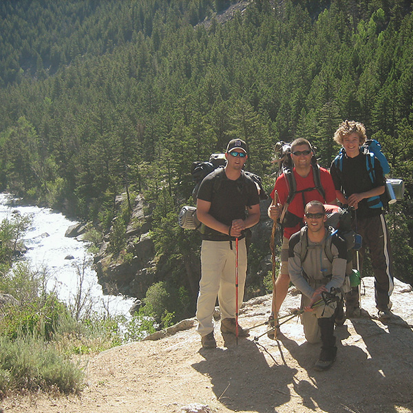 4 smiling young men with hiking poles standing on a rocky looking down on a forest with a stream running through it