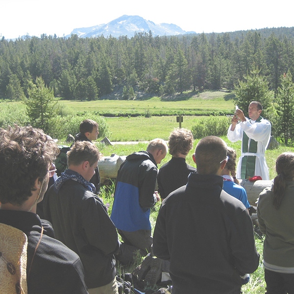 a priest blessing a communion wafer in front of a group of young people outside surrounded by trees and with mountains in the distance